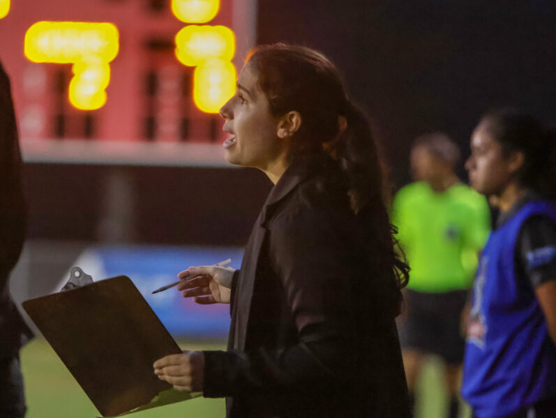 A female student assistant coach holds a clipboard and yells on the sidelines during a soccer game.